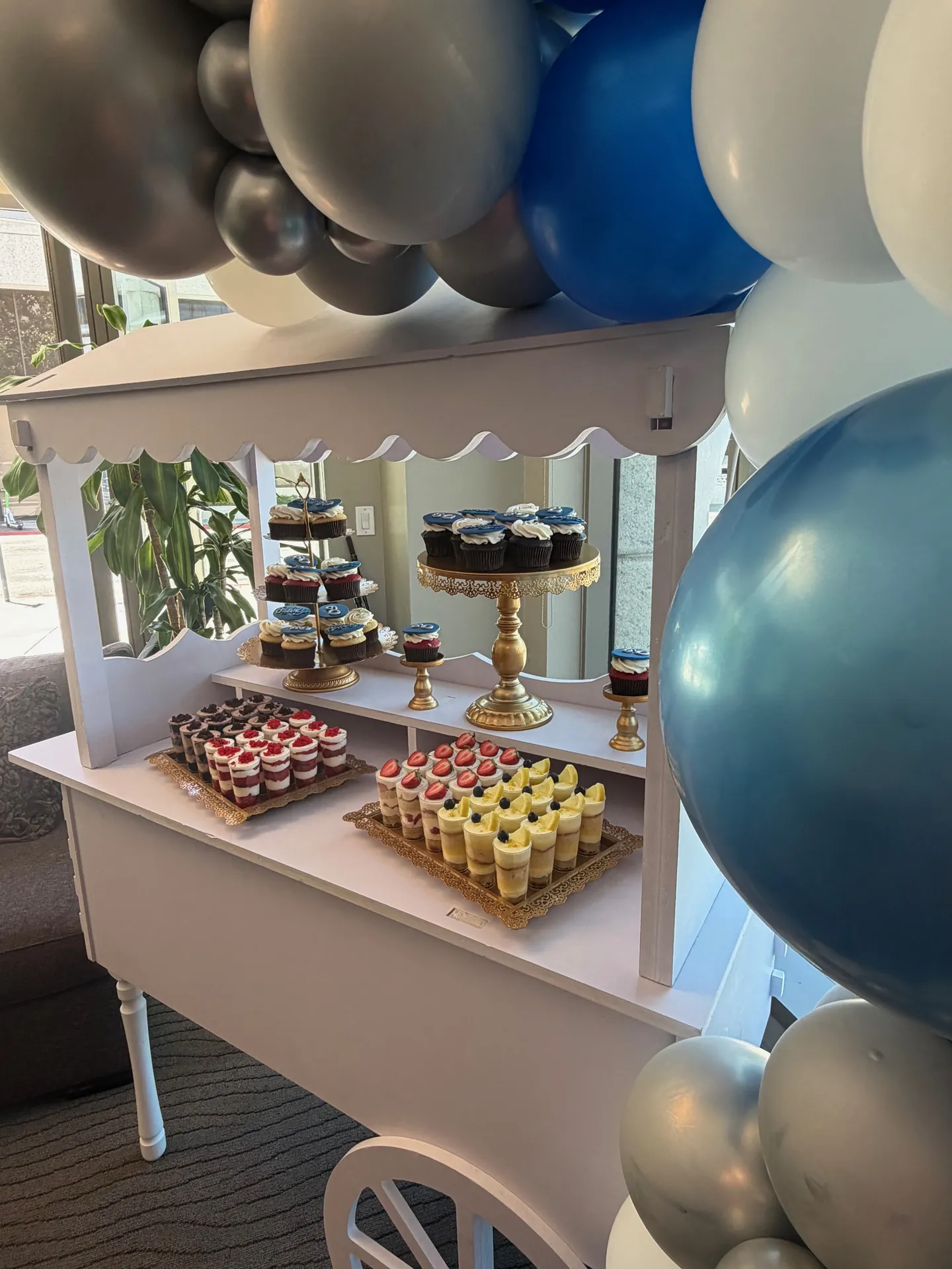 White dessert cart with cupcakes and dessert cups under blue and silver balloons