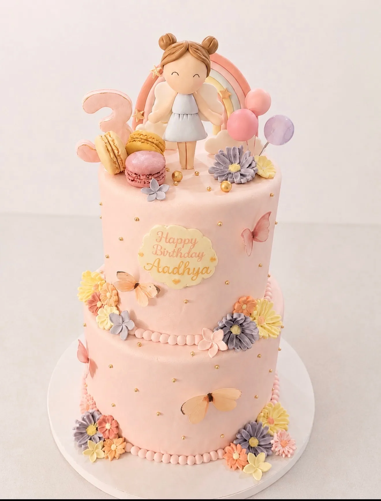Two-tier pink third birthday cake with fairy and rainbow topper, macarons, pastel flowers, butterflies, and gold pearls