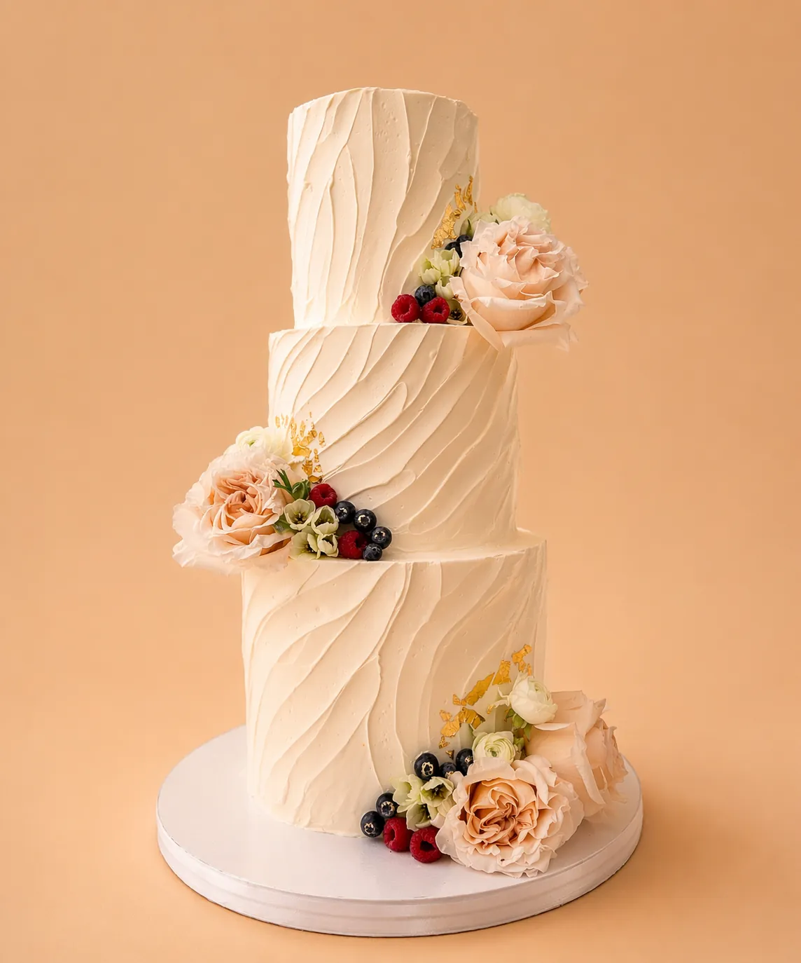 Three-tier wedding cake with textured buttercream, blush flowers, berries, and gold leaf