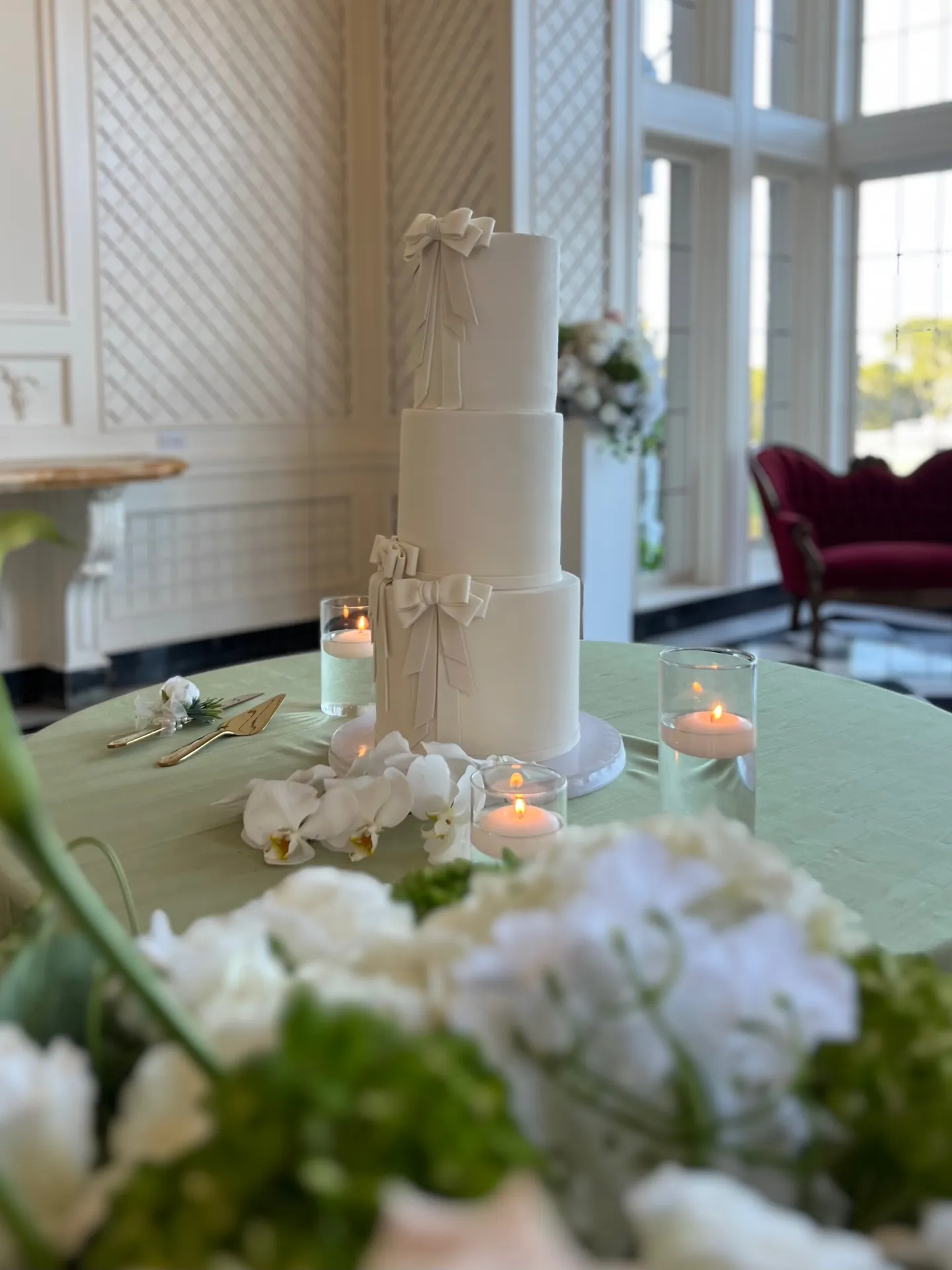 Three-tier white bow wedding cake displayed with flowers and candles at an event venue